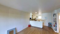 This single-floor space is designed with an open-concept layout that seamlessly connects the living area to the kitchen. The interior is finished in a clean, all-white palette, with paneled walls adding subtle texture to the living space. Light wood laminate flooring runs throughout, contributing warmth and continuity across the open floor plan.

The kitchen is positioned at the far end of the space and features a white peninsula counter that extends outward, providing additional workspace and serving as a natural divider between the kitchen and the living area. White cabinetry lines the upper walls of the kitchen, offering ample storage, while a refrigerator and basic appliances are visible, indicating a fully functional setup. Recessed lighting illuminates the kitchen area, complementing the natural light filtering in through a window.

The living area is spacious and open, with smooth white walls and a clean ceiling that enhance the sense of airiness throughout. The minimalist design approach keeps the space feeling uncluttered and versatile, making it well-suited for a variety of uses. Overall, the space presents a bright, functional, and understated aesthetic.