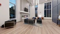 Located at 195 Dundas Street in London, Ontario, this contemporary amenity space offers a thoughtfully designed, single-floor layout that combines comfort, recreation, and functionality. The main lounge area makes an immediate impression with soaring double-height ceilings, floor-to-ceiling windows that flood the space with natural light, and rich views of the surrounding cityscape. A sleek wall-mounted linear fireplace framed in stone-effect tile serves as a striking focal point, complemented by a wall-mounted television above a clean, minimalist mantle. Comfortable seating arrangements with black leather sofas and accent chairs are set atop stylish area rugs over warm wood-tone luxury vinyl flooring, creating an inviting social atmosphere. Deep navy panel-clad accent walls add a bold, sophisticated contrast throughout the lounge corridor, which connects seamlessly to the various amenity zones. A fully appointed kitchen features crisp white cabinetry, quartz countertops, a subway tile backsplash, a double stainless steel sink, and a stainless steel dishwasher — ideal for hosting and entertaining. A dedicated fitness area provides ample room for exercise equipment, while a standout indoor golf simulator room, enclosed by glass partitions and featuring artificial turf, a projector, and a large screen, delivers a one-of-a-kind recreational experience. Throughout, the design maintains a cohesive modern aesthetic with clean lines, neutral tones, and high-quality finishes.