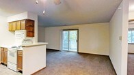 This single-floor space features an open-concept layout that integrates the living area with the kitchen. The living room is finished with wall-to-wall carpeting in a neutral tone, providing a comfortable and cohesive feel throughout. A sliding glass door serves as a focal point of the main living area, allowing natural light to filter into the space while offering direct access to an outdoor balcony enclosed with a decorative lattice railing. Views of surrounding greenery are visible through the glass door, adding a natural element to the interior ambiance.

The kitchen area is partially separated from the living space by a raised counter with a light-colored surface, creating a functional breakfast bar or pass-through. Dark wood upper cabinetry with open shelving provides storage and a warm contrast to the neutral walls. A pendant light fixture hangs from the ceiling above, adding a subtle decorative touch. The ceiling throughout features a textured finish. The overall atmosphere of the space is understated and practical, with an open feel enhanced by the connection to the outdoor balcony.