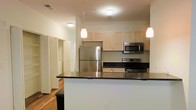 This single-floor residence offers a thoughtfully designed layout with a clean, modern aesthetic. The kitchen serves as a central highlight, featuring stainless steel appliances including a full-size refrigerator, electric range, and over-the-range microwave. Rich dark granite countertops provide generous workspace and contrast beautifully against the light maple cabinetry above. A peninsula-style breakfast bar extends the counter space and creates a natural divide between the kitchen and the adjoining living area, making it ideal for casual dining or entertaining. Pendant lighting hangs above the peninsula, adding a warm, inviting ambiance to the space. Warm hardwood flooring flows throughout, contributing to a cohesive and polished look. The interior benefits from recessed and flush-mount ceiling lighting, ensuring the space is well-lit throughout the day. Beyond the kitchen, a hallway leads to additional rooms, offering a functional and organized floor plan. The overall design balances practicality with modern style, making this a comfortable and visually appealing living space.
