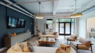 This thoughtfully designed single-floor commercial space blends industrial-chic architecture with contemporary furnishings to create a dynamic and functional environment. The main lounge area serves as the heart of the space, featuring exposed white-painted ductwork and piping along the ceiling, bold navy accent walls, and warm wood-look flooring. Generous seating arrangements with neutral-toned sofas, patterned throw pillows, and a live-edge coffee table create an inviting atmosphere, while large glass-paneled entry doors and windows flood the area with natural light and offer views of the surrounding greenery. A wall-mounted flat-screen TV and a sideboard complete the lounge's practical appeal. Adjacent to the lounge, a sleek kitchenette is outfitted with dark cabinetry, granite-style countertops, a stainless steel sink, a beverage refrigerator, and dramatic vertical tile backsplash with open shelving. A dedicated conference room features a substantial solid wood table, modern globe pendant lighting, curated wall art, and a bright window. Additional flexible workspace areas include semi-private rooms accessed via sliding barn-style doors, accented by statement drum pendant lights and casual seating nooks, making this space well-suited for collaborative and focused work alike.