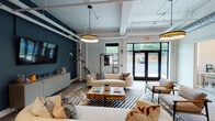 This thoughtfully designed single-floor commercial space blends industrial-chic architecture with contemporary furnishings to create a dynamic and functional environment. The main lounge area serves as the heart of the space, featuring exposed white-painted ductwork and piping along the ceiling, bold navy accent walls, and warm wood-look flooring. Generous seating arrangements with neutral-toned sofas, patterned throw pillows, and a live-edge coffee table create an inviting atmosphere, while large glass-paneled entry doors and windows flood the area with natural light and offer views of the surrounding greenery. A wall-mounted flat-screen TV and a sideboard complete the lounge's practical appeal. Adjacent to the lounge, a sleek kitchenette is outfitted with dark cabinetry, granite-style countertops, a stainless steel sink, a beverage refrigerator, and dramatic vertical tile backsplash with open shelving. A dedicated conference room features a substantial solid wood table, modern globe pendant lighting, curated wall art, and a bright window. Additional flexible workspace areas include semi-private rooms accessed via sliding barn-style doors, accented by statement drum pendant lights and casual seating nooks, making this space well-suited for collaborative and focused work alike.