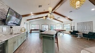 Located at 2330 SW Williston Rd in Gainesville, Florida, this single-story clubhouse is designed to serve as a social and recreational hub. The space is defined by its dramatic vaulted ceilings adorned with rustic exposed wood beams, which lend warmth and architectural character throughout. Wood-look tile flooring runs continuously across the entire space, providing a cohesive and durable foundation.

The main gathering area features an open-concept layout centered around a granite-topped island with bar seating and a curved wet bar finished in sage green cabinetry. The bar area includes a sink, wine refrigerator, and a textured tile accent wall with a mounted television. Decorative pendant lighting in woven and geometric gold styles hangs from the ceiling, adding an eclectic, modern touch.

A game room section accommodates a full-size billiard table and a shuffleboard table, while an adjacent lounge area offers plush seating, velvet accent chairs, and a wall-mounted fireplace framed in stacked stone. Large windows throughout the space provide abundant natural light and views of the lush surrounding greenery. A cozy nook features an oversized Scrabble board and curved bench seating, adding a playful, interactive element to the overall design.