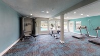 This fitness center offers a comprehensive workout environment designed to support a wide range of exercise routines. The space is finished with a vibrant teal accent wall that adds energy to the room, complemented by neutral gray walls, white trim, and a textured ceiling with recessed lighting throughout. The patterned carpet flooring in shades of blue, gray, and brown provides both comfort and a modern aesthetic.

The layout is open and well-organized, with structural columns defining distinct workout zones without fully dividing the space. The cardio area features treadmills and an elliptical trainer positioned near large windows and a glass door that open to views of an outdoor pool and patio, allowing natural light to fill the room. A dedicated strength training section houses a multi-function cable machine, a weight bench, and various resistance equipment. A functional fitness corner is stocked with stability balls in multiple sizes, resistance bands, and additional accessories, making it suitable for stretching and dynamic exercises.

The space benefits from ample natural light streaming through multiple windows on multiple walls, creating a bright and motivating atmosphere. An adjacent area visible through an open passage appears to offer additional seating or lounge space, adding versatility to the overall facility.