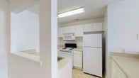 This single-floor residential unit offers a clean and straightforward layout designed for comfortable everyday living. The living area is finished with neutral-toned carpeting and crisp white walls, creating a blank canvas that is easy to personalize. Multiple electrical outlets are evenly distributed along the walls, providing practical convenience throughout the space. A doorway visible from the living area leads to an adjacent room, suggesting a well-connected floor plan. The kitchen is efficiently arranged and outfitted with white cabinetry that extends to the upper walls, maximizing storage capacity. A full-size refrigerator, an electric range, and an over-the-range microwave are included, offering all the essentials for meal preparation. Light-toned countertops and tile flooring contribute to the kitchen's clean, cohesive aesthetic, while a built-in sink adds further functionality. Overhead fluorescent lighting ensures the kitchen is well-illuminated. A textured ceiling finish is consistent throughout the space. Overall, the unit presents a tidy, move-in-ready environment with a practical design suited for a range of occupants.