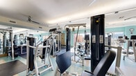 Located at 1333 Old Spanish Trail in Houston, TX, this fitness center offers a spacious, single-floor layout designed to accommodate a comprehensive range of workout activities. The facility is divided into two primary areas: a main strength and cardio training floor and a dedicated cycling studio.

The main workout area features light teal accent walls, rubberized flooring, and abundant natural light streaming through large sliding glass doors that open to views of the surrounding cityscape. Ceiling fans throughout help maintain a comfortable environment. The space is outfitted with a full suite of Magnum strength machines, free weights including a rack of dumbbells, benches, and cardio equipment such as treadmills and rowing machines. Wall-mounted TVs and full-length mirrors add to the functional design, while framed artwork contributes a polished aesthetic.

The cycling studio is a bright, dedicated room featuring warm bamboo hardwood flooring, floor-to-ceiling mirrors along one wall, and multiple ceiling fans. A full row of stationary spin bikes, stability balls, and wall-mounted flat-screen TVs make this space ideal for group fitness classes or individual cycling sessions.

Together, the two areas form a well-rounded fitness facility that balances functionality with a clean, modern design.