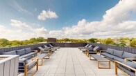 This expansive rooftop terrace offers a well-designed outdoor retreat elevated above the surrounding neighborhood. The terrace is finished with large-format light-toned pavers that lend a clean, contemporary aesthetic throughout the open-air space. A sleek, dark corrugated metal parapet wall frames the perimeter, providing a defined boundary while allowing unobstructed panoramic views of lush treetops and the broader cityscape beyond.

The terrace is thoughtfully furnished with multiple seating arrangements, including teak-framed lounge chairs with deep grey cushions and matching low coffee tables, creating inviting conversation areas across the space. A generous collection of reclining sun loungers with teak frames is arranged to maximize sun exposure, ideal for relaxation. Tucked along one section of the terrace is a dedicated lounge nook with additional seating and accent greenery, adding a softer, more intimate atmosphere. A centrally positioned built-in outdoor kitchen or grill station provides amenities for outdoor entertaining.

Access to the terrace is provided through a glass-paneled door set within the dark corrugated metal rooftop structure, which also features skylight elements. The open-sky setting floods the entire terrace with abundant natural light throughout the day, making it a highly functional and visually striking outdoor amenity space.
