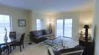 This single-floor space centers around a well-appointed living room designed for both comfort and functionality. The room is finished with neutral-toned walls and carpeted flooring, contributing to a warm and inviting atmosphere. Crown molding runs along the ceiling perimeter, adding a subtle architectural detail that elevates the overall aesthetic.

Natural light is a defining feature of the living room, with multiple large windows fitted with horizontal blinds allowing for adjustable brightness. A expansive sliding glass door or wide window unit along one wall opens the space visually to an outdoor view, suggesting an elevated vantage point with surrounding exterior scenery visible beyond a railing.

The living area is furnished with a dark leather sofa accented by striped throw pillows, paired with a sleek rectangular coffee table resting on a geometric patterned area rug. Additional seating is positioned nearby, and a tall floor lamp with a tripod base provides supplemental ambient lighting. A framed artwork piece on the wall and a small side table complete the thoughtful arrangement, making the space feel curated and livable.