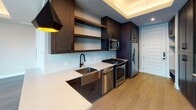 This single-floor residence showcases a thoughtfully designed interior defined by contemporary finishes and a cohesive dark-and-light aesthetic throughout. The kitchen is a functional centerpiece, outfitted with rich dark wood cabinetry, open floating shelves, a textured tile backsplash, white quartz countertops, and a stainless steel farmhouse sink. A full suite of stainless steel appliances — including a gas range, dishwasher, built-in microwave, and refrigerator — provides a well-equipped cooking environment. Recessed tray ceilings with integrated ambient lighting add architectural depth to this area.

The main living space is expansive and airy, highlighted by a dramatic floor-to-ceiling glass wall with dark steel framing that floods the interior with natural light and offers direct views of an outdoor patio. A ceiling fan with integrated lighting is mounted within the tray ceiling, enhancing comfort and airflow. Built-in dark wood shelving and cabinetry along one wall provide generous storage and display space.

The bathroom is finished with warm-toned large-format floor tiles, dark wood vanity cabinetry, white quartz countertops, pendant lighting, a wide mirror, and a full tub-shower combination with detailed tile surround. Light wood flooring runs consistently throughout the living areas, unifying the overall design.