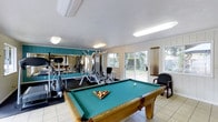 This single-floor amenity space seamlessly combines recreational and fitness functions into one well-appointed room. At the center sits a full-size pool table with a classic green felt surface and warm wood frame, making it an immediate focal point for leisure and social activity. The billiards area is complemented by light-toned tile flooring that extends throughout the space, lending a clean and open feel.

The fitness area occupies one side of the room and is outfitted with a multi-station strength training machine, an elliptical trainer, and additional exercise equipment. A large wall-mounted mirror runs the length of the workout zone, visually expanding the space and providing exercisers with a full view during their routines. Teal-painted wall paneling adds a bold accent that energizes the fitness area.

Natural light floods the room through multiple large sliding glass doors and windows that face a lush outdoor area, creating an inviting, airy atmosphere throughout. The sliding doors also offer convenient access to the exterior. Overhead fluorescent lighting ensures the space remains well-lit at all hours. The room's dual-purpose layout makes it a highly functional space suited for both active workouts and casual recreation.