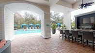 This single-story covered outdoor pavilion offers a well-appointed resort-style amenity space designed for relaxation and entertainment. The structure features a generous covered patio with a high ceiling, recessed lighting, and ceiling fans, creating a comfortable, shaded environment that opens directly to the surrounding pool area.

The pavilion is anchored by a sleek outdoor kitchen and bar, complete with dark granite countertops and wicker bar seating for multiple guests. Adjacent to the bar, a built-in fireplace topped with a mounted flat-screen television serves as a focal point, with a curved wicker sectional sofa with bold rust-colored cushions arranged in front for comfortable viewing and gathering.

A separate dining area features a glass-top table surrounded by wicker chairs, positioned near large windows that connect the covered patio to an interior space. Lush tropical potted plants are strategically placed throughout, adding a verdant, resort-like atmosphere.

The entire patio is finished with classic brick pavers that extend to the pool deck, where striped blue cabanas and lounge chairs are arranged alongside a shimmering pool. Mature oak and palm trees frame the backdrop, enhancing the natural, relaxed ambiance of the space.