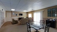 This single-floor space functions as a comfortable common area, thoughtfully arranged to accommodate both lounging and dining. The room features a drop tile ceiling with recessed lighting panels that provide consistent, even illumination throughout. A large sliding glass door fitted with vertical blinds allows natural light to fill the space, contributing to a bright and welcoming atmosphere.

The living area is furnished with a sectional sofa and an armchair in neutral tones, arranged around a wall-mounted flat-screen television. A compact sideboard sits beneath the TV, offering additional storage. Framed artwork on the warm beige walls adds a modest decorative touch, complementing the subdued color palette of the space.

Adjacent to the seating area, a glass-top dining table surrounded by dark-framed chairs with cushioned seats provides a defined area for meals or gatherings. The flooring transitions between tile near the entrance and patterned carpet in the lounge zone, subtly delineating the functional areas. Overall, the space presents a clean, functional, and moderately appointed environment designed for everyday communal use.