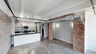 This single-floor loft seamlessly blends industrial character with modern finishes, creating a striking and functional living space. Throughout the unit, polished concrete floors provide a sleek, durable foundation, while exposed brick walls add warmth and raw texture that speak to the building's historic roots. Structural ceiling beams and exposed cylindrical ductwork run overhead, reinforcing the loft's industrial aesthetic.

The open-concept living area is generously proportioned, benefiting from large windows that flood the space with natural light and offer views of the surrounding cityscape. A ceiling fan serves the main living zone, enhancing comfort and airflow.

The kitchen is well-appointed with white shaker cabinetry, dark granite countertops, and a full suite of stainless steel appliances, including a range, microwave, and refrigerator. A large kitchen island with an undermount sink and a commercial-style faucet serves as a central hub for meal preparation and casual dining. Open shelving adds both practicality and visual interest.

The bathroom continues the industrial-modern theme, featuring a sleek floating double vanity, large windows framed by exposed brick, and an abundance of natural light with direct city views. Track lighting and recessed fixtures throughout the space ensure well-lit interiors at any time of day.