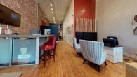 This newly renovated single-floor space sits prominently in the heart of an arts district, offering a thoughtfully designed interior that balances historic architectural character with contemporary style. Large street-facing windows flood the front of the space with natural light and provide open views of the surrounding neighborhood. A built-in window bench with dark upholstery and colorful accent pillows creates an inviting seating nook along the storefront, enhancing the welcoming atmosphere.

Exposed brick walls run throughout much of the interior, lending warmth and texture that complement the original hardwood floors underfoot. The space features soaring ceilings with visible industrial-style track lighting, adding both drama and practical illumination. Bold accent walls in deep red and teal punctuate the open layout, while curated artwork—including large-scale abstract paintings and sculptural wall pieces—reinforces the arts-forward identity of the location.

Distinct functional zones are arranged throughout the open floor plan, including a comfortable lounge area with plush seating, a workstation area, and a bar-height counter with red leather stools. A private office is accessible via a glass-paneled door near the entrance. The overall ambiance is creative, polished, and energetic.