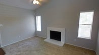 This single-story home in Arlington, Texas offers a welcoming living space defined by its vaulted ceilings, which lend an open, airy feel to the main living area. The room's most prominent feature is a white-painted brick fireplace with a raised hearth, serving as a natural focal point and adding a classic, traditional character to the space. Neutral gray walls pair seamlessly with the beige wall-to-wall carpeting, creating a clean and versatile backdrop suitable for a variety of interior styles. A ceiling fan is centrally mounted, providing both functional air circulation and a modest decorative element that complements the room's understated aesthetic. Natural light enters through a vertically oriented window dressed with horizontal blinds, offering privacy while still brightening the space. The combination of vaulted ceilings and open wall space gives the living area a generously proportioned feel. Overall, the space presents a straightforward, functional layout with timeless design elements, making it a practical and comfortable living environment in the heart of the Dallas–Fort Worth metroplex.