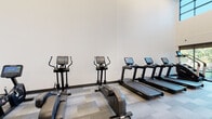 This fitness center is designed to support a full range of workout routines within a thoughtfully organized, single-floor layout. The space is divided into distinct zones for cardio and strength training, each outfitted with modern equipment. The cardio area features multiple treadmills, elliptical trainers, a stair climber, and a rowing machine, all equipped with digital display consoles. The strength training section offers a variety of cable machines, weight benches, and resistance equipment arranged to allow ample circulation between stations.

The interior features a neutral color palette of whites and grays, with patterned carpet tile flooring throughout. Recessed and flush-panel lighting provides consistent illumination across the space. Columns with warm wood accent detailing add architectural interest to the strength training area. Floor-to-ceiling windows along one wall allow natural light to enter and offer views of the surrounding outdoor greenery, enhancing the overall atmosphere. A designated entry area includes posted fitness center rules, a bench, and clearly marked emergency exits, reflecting attention to both function and safety standards.