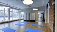 This single-floor wellness studio is thoughtfully designed to support a range of fitness and mindfulness activities. The main exercise area features warm hardwood flooring and is currently arranged with multiple blue yoga mats, making it well-suited for yoga, stretching, or group fitness classes. Large windows along one wall flood the space with abundant natural light and offer views of the surrounding outdoor landscape, creating a bright and inviting atmosphere conducive to focused practice.

Overhead, circular pendant light fixtures provide even, ambient illumination throughout the room, complementing the natural light. The ceiling incorporates a raised tray detail that adds architectural dimension to the space. Neutral gray walls contribute a calm, clean aesthetic throughout.

A folding shoji-style room divider offers flexible partitioning within the studio, allowing the space to be reconfigured as needed. A full-length mirror along one wall visually expands the room and provides a clear sightline into an adjacent fitness equipment area, which is visible in the reflection and appears to house cardio and strength-training machines. Together, these elements make this a versatile, well-appointed facility for both group and individual fitness activities.