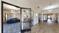 Located at 15600 Galaxie Ave in Apple Valley, Minnesota, this single-floor community amenity center offers a generous and well-appointed space designed for recreation, relaxation, and socializing. The centerpiece is a striking indoor swimming pool, enclosed within a soaring vaulted ceiling of warm wood, surrounded by stone block walls and softly lit sconces that create an inviting aquatic retreat. Connected to the pool area are wide, tiled hallways with clean white walls, providing easy navigation throughout the facility. The expansive lounge and gathering area serves as the social heart of the space, featuring plush navy leather seating arranged around a classic brick fireplace, complemented by elegant chandeliers, ceiling fans, and large windows that allow abundant natural light and views of the surrounding landscape. Multiple dining areas offer ample seating at wooden tables and chairs, one highlighted by a decorative mirror and wall sconces for an added touch of warmth. Glass-partitioned entryways with modern black metal framing create a transparent, open feel as one moves between areas. The fitness room is well-equipped with treadmills, a multi-station weight machine, and stationary bikes, with wall-to-wall mirrors enhancing the sense of space. Carpet and tile flooring throughout provide comfort and practicality, making this a comprehensive and welcoming community facility.