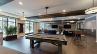 This single-story clubhouse offers a well-appointed community gathering space designed with a modern-rustic aesthetic. Reclaimed wood beam accents, rich hardwood-style flooring, and a mix of stone and wood wall treatments create a warm yet contemporary atmosphere throughout. The open layout flows seamlessly between distinct activity zones, each thoughtfully furnished and equipped.

The main lounge area features large windows overlooking an outdoor pool courtyard, filling the space with natural light. A plush navy sectional, leather accent chairs, and a glass coffee table anchor this inviting seating area. Nearby, a stone-clad linear fireplace with a mounted flat-screen TV provides a cozy focal point, complemented by additional lounge seating.

A dedicated game room includes a full-size billiard table, ping-pong table, and a classic arcade machine, catering to a range of recreational interests. The dining and entertaining area offers multiple round tables with upholstered seating alongside a bar counter with marble-look surfaces, pendant lighting, and bar stools.

A reclaimed wood accent wall with a large mounted TV defines a secondary media lounge. Glass-walled rooms provide flexible enclosed spaces visible from the main corridor, while a rustic barn door adds a charming architectural detail. The overall ambiance is social, stylish, and welcoming.