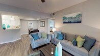 This well-appointed single-floor residence offers a thoughtfully designed open-concept layout that promotes a natural flow between living spaces. The living room serves as the heart of the home, anchored by plush gray sofas and accent chairs adorned with teal and green throw pillows, creating a fresh, inviting color palette. Warm hardwood-style flooring runs throughout, adding a sense of continuity and warmth to the space.

A vaulted ceiling detail elevates the living area, adding architectural interest and a sense of openness. A ceiling fan provides both functionality and a finishing touch to the overhead space. A bold teal abstract canvas on the wall ties together the room's coastal-inspired color scheme.

The dining area is positioned just off the living room, separated by subtle architectural framing, and features a dark-toned dining table with matching chairs, accented by a framed wall piece. A partially visible bedroom beyond the living space is bright and warmly lit, suggesting a comfortable and cohesive design throughout the residence. The neutral gray wall tones throughout the home provide a versatile backdrop that complements the vibrant accent colors and modern furnishings, resulting in a space that feels both contemporary and welcoming.