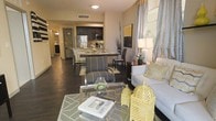 This single-floor residence showcases an open-concept design that integrates the living, dining, and kitchen spaces into one cohesive, flowing layout. The living area is anchored by a plush white sofa accented with gold and green throw pillows, paired with a mirrored glass coffee table that adds a touch of elegance. Decorative elements, including a gold birdcage accent piece and patterned area rug, contribute to the space's curated aesthetic. Floor-to-ceiling curtains in a neutral tone with subtle patterns frame the windows, allowing natural light to filter through.

The dining area transitions naturally from the living space, featuring a sleek bar-height table with upholstered gray chairs, creating an ideal setting for casual dining. The kitchen is equipped with stainless steel appliances, dark cabinetry, and a light-toned countertop, balancing contrast and functionality. Rich, dark hardwood flooring runs throughout, providing a warm and grounding foundation. The overall ambiance is modern and sophisticated, combining neutral tones with gold accents for a refined, contemporary feel.
