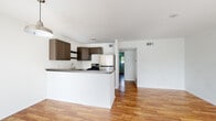 This single-floor unit offers a clean, functional layout designed for comfortable everyday living. Warm hardwood flooring runs continuously throughout the space, lending a cohesive and inviting feel to every room. The open-concept living and kitchen area forms the heart of the unit, where a peninsula-style kitchen seamlessly connects to the living space. The kitchen is well-appointed with taupe shaker-style cabinetry, open shelving, stainless steel appliances including a gas range and refrigerator, and ample counter space with dual-tone countertops. The living area is generously sized and bathed in natural light through large front-facing windows dressed with sheer curtains, with a front entry door providing direct outdoor access to what appears to be a shared balcony or walkway. The bedrooms feature ceiling fans, hardwood flooring, and windows overlooking greenery outside. One bedroom includes a walk-in closet, adding practical storage. The bathroom is neatly appointed with a full tub-shower combination, white surround, a vanity with matching cabinetry, and wood flooring. Throughout, the unit maintains a neutral white palette, creating a bright and airy atmosphere well-suited to a variety of interior styles.