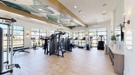 This single-floor fitness center offers a thoughtfully designed workout environment that balances functionality with aesthetic appeal. The space is flooded with natural light through expansive floor-to-ceiling black-framed windows that line the perimeter, providing open views of the surrounding outdoor landscape, including palm trees and greenery that reinforce a bright, resort-like atmosphere.

The ceiling is a standout architectural feature, adorned with a large-scale tropical botanical mural depicting oversized leaves in soft greens and whites. Exposed wood beams and a ceiling fan complement the design, adding warmth and a relaxed, coastal character. Pendant lighting and wall sconces provide additional ambient illumination throughout.

Light hardwood-style flooring runs throughout the entire space, contributing to a clean and modern aesthetic. The fitness floor is comprehensively outfitted with a multi-station cable weight machine, a free weights rack with dumbbells and an adjustable bench, a power rack, treadmills with individual digital displays, stationary cycling bikes, and stair climbers.

A dedicated entryway area features a wall-mounted flat-screen TV, open storage shelving, and a decorative console, creating a welcoming transition into the main workout floor. Full-length mirrors along one wall enhance the sense of space and support training visibility. Glass-paneled interior doors connect areas seamlessly, reinforcing the open, airy feel of the facility.