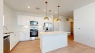 This single-floor residence offers a well-organized layout encompassing two bedrooms, two bathrooms, a dedicated laundry room, and two closets. The open-concept kitchen and living area serves as the heart of the home, featuring white shaker cabinetry, a full suite of stainless steel appliances including a French door refrigerator, electric range, and microwave. A central island with a white countertop provides additional prep space and casual seating, while two gold-toned pendant lights add a warm, modern accent. Light wood-look flooring flows throughout the main living areas and bathrooms, contributing to a bright, cohesive aesthetic.

The bedrooms are finished with neutral carpet and receive natural light through window blinds. The primary bedroom connects directly to a bathroom equipped with a white vanity, ample cabinetry, and an adjacent walk-in closet with built-in shelving. The dedicated laundry room accommodates a full-size washer and dryer with overhead shelving for storage. Throughout the unit, crisp white walls, recessed lighting, and a clean, minimalist design create a consistent and functional living environment.