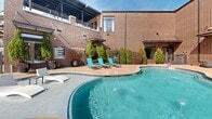 This contemporary residential property boasts an impressive outdoor amenity space centered around a stunning resort-style pool. The pool area is expansive, featuring a uniquely shaped design with multiple sections, including a curved edge and built-in seating areas. The pool is surrounded by a spacious concrete deck dotted with numerous turquoise lounge chairs, creating a cohesive and inviting aesthetic.

The architecture of the adjacent building is notable, with a brick exterior and large, multi-paned windows that allow natural light to flood the interior spaces. A covered patio area extends from the building, providing shade and seamlessly connecting indoor and outdoor living areas. Architectural elements such as pergolas and balconies add visual interest and functionality to the structure.

Landscaping around the pool area includes well-manicured shrubs and trees, adding a touch of greenery to the urban setting. The overall design creates a harmonious blend of modern architectural elements with resort-like amenities, offering residents a luxurious and relaxing environment. The space appears well-maintained and designed for both aesthetics and practical use, making it an attractive feature for the residential complex.