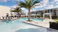 This stunning resort-style pool area delivers a true vacation atmosphere right at home. The centerpiece is a sprawling, crystal-clear turquoise pool with a distinctive design that incorporates a central island planter, home to a majestic palm tree rising dramatically from the water's edge. The pool deck is finished in crisp, light-toned tile that reflects the Florida sunshine beautifully. Surrounding the pool, generous lounge areas are outfitted with comfortable chaise loungers and a series of elegant white fabric cabanas and market umbrellas, providing shaded retreats for relaxation throughout the day. Lush tropical landscaping, including multiple towering palm trees, reinforces the resort ambiance and adds natural beauty to the space. The modern residential buildings in the background feature a clean, contemporary architectural style with yellow and white façades, balconies, and large windows. The open-sky setting allows abundant natural light to flood the area, creating a bright, airy, and inviting outdoor environment. Whether lounging in the sun or seeking shade under a private cabana, this thoughtfully designed amenity space offers an exceptional outdoor living experience with a polished, upscale aesthetic.
