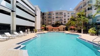 This outdoor amenity area centers around a generously sized swimming pool with clear, blue water, creating a resort-like atmosphere within a modern residential complex. The pool deck is well-appointed with lounge chairs arranged along the perimeter, offering plenty of space for sunbathing and relaxation. A centrally located cabana structure provides shaded seating areas beneath vibrant red umbrellas, serving as a social hub within the outdoor space. Lush tropical palms and manicured greenery are strategically placed around the deck, enhancing the leisure ambiance and adding a warm, subtropical character. The surrounding multi-story building features rows of private balconies that overlook the pool courtyard, while an adjacent parking structure completes the enclosed setting. The open-sky courtyard layout allows abundant natural sunlight to illuminate the space throughout the day, contributing to its bright and inviting atmosphere. The combination of contemporary architecture, well-maintained hardscaping, and tropical landscaping gives this outdoor area a polished, amenity-rich character well-suited for active outdoor enjoyment and casual social gatherings.