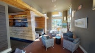 This single-story clubhouse is thoughtfully designed to serve as a dynamic community gathering space, blending modern aesthetics with warm, natural materials. The interior is anchored by a striking natural wood-clad feature wall with horizontal planking that defines the lounge area, where plush upholstered chairs are arranged around a central coffee table atop a rich, patterned carpet. Soaring ceilings with floor-to-ceiling windows invite generous natural light throughout and frame picturesque outdoor views of surrounding greenery and the surrounding landscape.

A dedicated media nook features a large wall-mounted flat-screen television, wood-paneled accent wall, and a leather loveseat — ideal for relaxed viewing. The casual dining area offers bistro-style tables with stools positioned beside tall windows overlooking palm trees, accented by a decorative wall clock and contemporary sconce lighting.

The fully equipped kitchen is outfitted with dark cabinetry, stone-look countertops, stainless steel appliances including a refrigerator, range, and dishwasher, and a mosaic tile backsplash beneath a slatted wood ceiling detail. A billiards table anchors the central entertainment area, completing the clubhouse's diverse amenity offerings. The main entrance features warm wood-framed glass doors, adding an inviting, polished finish to this versatile community space.