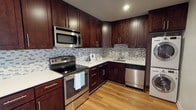 This single-floor space is thoughtfully designed with a focus on functionality and modern style. The kitchen serves as a standout feature, showcasing rich, dark wood cabinetry that extends to the ceiling, maximizing storage while creating a bold, elegant aesthetic. The cabinetry is complemented by light-toned countertops that provide a striking contrast, and a mosaic tile backsplash in soft blues and neutrals adds a refined decorative touch throughout the cooking area.

The kitchen is fully equipped with a suite of stainless steel appliances, including an electric range with a built-in over-the-range microwave, a dishwasher, and an in-unit washer, highlighting the space's emphasis on convenience. The undermount sink is positioned beneath the backsplash and integrated seamlessly into the counter layout, while recessed lighting overhead ensures the workspace is well-lit.

Warm hardwood flooring runs throughout, lending the space a welcoming and cohesive feel. The layout is efficient and well-organized, with an L-shaped kitchen configuration that promotes ease of movement. Overall, the space balances practical design with contemporary finishes, making it both visually appealing and highly functional for everyday living.