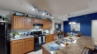 This single-floor residence combines coastal-inspired design with practical comfort throughout its thoughtfully arranged layout. The living room serves as a focal point, anchored by a striking deep-blue accent wall adorned with nautical artwork, complemented by hardwood-style flooring, crown molding, a ceiling fan, and a sliding glass door that invites natural light and outdoor views. The open-concept kitchen is well-equipped with warm honey-toned cabinetry reaching to the ceiling, granite-style countertops, black appliances including a range and full-size refrigerator, a double stainless steel sink, and track lighting overhead. The kitchen flows seamlessly into a dedicated dining area illuminated by a chandelier, creating an inviting space for entertaining. The bedrooms offer a calm retreat, featuring plush carpeting, ceiling fans, and generous natural light through large windows with blinds and drapery. The primary bathroom includes a walk-in glass-enclosed shower with tile flooring and neutral tones throughout. A dedicated laundry room houses a full-size washer and dryer with built-in shelving above, adding everyday convenience. The cohesive blue-and-white color palette ties the entire residence together, creating a relaxed yet refined coastal ambiance.