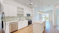 This thoughtfully designed one-bedroom, one-bathroom residence offers a bright and modern living experience across a single floor. The open-concept kitchen and living area serves as the heart of the home, featuring warm light wood flooring, recessed lighting, and elegant brass-accented pendant lights that hang above a generous white kitchen island with built-in shelving. The kitchen is outfitted with crisp white shaker cabinetry, gold hardware, a soft green subway tile backsplash, stainless steel appliances including a refrigerator, range, microwave, and dishwasher, and a deep undermount sink with a flexible faucet. The living area extends toward a sliding glass door that opens to an outdoor view of a landscaped, fenced green space adorned with a vibrant floral mural. The bedroom is a comfortable retreat with plush carpet flooring, a ceiling fan, and a window that brings in natural light. Two closets provide ample storage, including a spacious walk-in closet with custom white shelving and gold hanging rods. The full bathroom is clean and well-appointed with a wide vanity, illuminated mirror, white cabinetry with gold hardware, and tile flooring. A stacked washer and dryer unit is conveniently integrated near the bathroom and closet area.