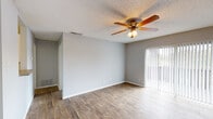 This single-story space is thoughtfully laid out to maximize functionality across its living areas. The main living room is a standout feature, showcasing durable wood-look flooring that extends throughout the primary living areas, neutral gray walls, and a ceiling fan with integrated lighting that promotes airflow and comfort. Vertical blinds frame a sliding glass door, allowing natural light to filter in while providing access to the outdoors.

The entryway transitions seamlessly from tile flooring into the wood-look flooring of the main living space, with a pendant light overhead and a nearby kitchen that is visible from the entry. The galley-style kitchen is well-equipped, featuring white cabinetry, dark countertops, a stainless steel sink, and black appliances including an electric range, refrigerator, and dishwasher. Light tile flooring completes the kitchen's clean aesthetic.

The bedroom offers plush carpeting underfoot and a window fitted with horizontal blinds, providing natural light and outdoor views. The bathroom includes a full tub-shower combination with white surrounds and tile flooring, maintaining a clean and functional design. Textured ceilings are consistent throughout the space.