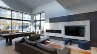 This contemporary single-floor space exemplifies modern design with a refined, minimalist aesthetic. The living area commands attention with its dramatically high ceilings and expansive floor-to-ceiling windows framed in dark metal, flooding the interior with natural light and offering open views of the surrounding urban environment. The centerpiece of the room is a striking fireplace wall clad in large-format marble-effect stone panels, paired with a sleek linear gas fireplace and an integrated flat-screen display, creating a bold and sophisticated focal point. Neutral tones throughout — from the light tile flooring to the crisp white walls and ceiling — provide a calm, gallery-like backdrop that allows key design elements to shine. The living area is anchored by a generously sized sectional sofa in rich charcoal leather, complemented by a transparent glass coffee table and an accent ottoman in deep blue velvet. A games or work table is visible near the windowed corner, adding a functional yet stylish touch. The interplay of natural light, high-end materials, and carefully curated furnishings gives the space a polished, upscale ambiance that is both welcoming and architecturally impressive.
