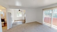 This single-floor unit offers a clean, functional layout with a neutral color palette throughout. The living area features plush carpeting and connects openly to an adjoining room, where a ceiling fan and a bright window allow natural light to flow freely. A sliding glass door in the living space leads to the outdoors and is fitted with vertical blinds, while a wall-mounted air conditioning unit provides climate control. The entry features a classic six-panel white door with a seamless transition between wood-look flooring and carpet. The kitchen is efficiently designed with white cabinetry, black hardware, white countertops, a stainless steel sink, and a window above the counter that brings in natural light. Multiple bedrooms offer comfortable carpeted floors, ample wall space, and windows that frame outdoor greenery. One bedroom includes a closet, adding practical storage. The bathroom is well-appointed with wood-look tile flooring, a dark-finished vanity with a white sink, a large mirror, and a full-size combination tub and shower with white tile surround and a small window for ventilation. Clean white walls and consistent finishes throughout create a cohesive and inviting atmosphere.