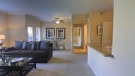 This single-floor residence offers a well-designed open-concept layout that seamlessly connects the main living areas. The living room is generously sized, featuring plush wall-to-wall carpeting, neutral beige walls, and a ceiling fan with integrated lighting that ensures year-round comfort. Natural light filters in through the windows, brightening the space and enhancing its warm, inviting ambiance. Framed artwork adorns the walls, adding a touch of character to the room.

The living area flows naturally into a dedicated dining space, which comfortably accommodates a full dining table and chairs — ideal for everyday meals or entertaining guests. Just beyond, the kitchen is highlighted by granite countertops on a raised breakfast bar, providing both functionality and an elevated aesthetic. The kitchen's layout suggests ample workspace and storage.

A central hallway extends from the main living area, leading to additional rooms beyond, offering a clear and practical floor plan. The consistent neutral color palette, carpeted flooring, and cohesive design elements throughout create a unified and comfortable atmosphere. Overall, this single-floor residence balances practicality with an approachable, homely style suited for comfortable everyday living.