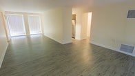 This single-floor space features a clean, modern aesthetic defined by smooth laminate wood-style flooring that extends throughout the open living area, lending warmth and visual continuity to the interior. The main living space is generously proportioned, with neutral cream-toned walls and white baseboards that create a bright, airy atmosphere. Natural light enters through a large window fitted with vertical blinds, illuminating the open floor plan effectively.

A wide passthrough opening connects the living area to the kitchen, which is visible from the main room and equipped with a stainless steel refrigerator. The kitchen's layout appears compact yet functional, with an additional adjacent area accessible through a secondary opening, suggesting further utility or storage space beyond.

The overall design is understated and practical, with minimal ornamentation allowing the architectural openness of the space to stand out. The smooth, uncluttered walls and consistent flooring throughout contribute to a cohesive and well-maintained environment. The single-floor layout ensures easy accessibility and straightforward navigation between areas, making the space both efficient and comfortable for everyday use.