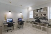 Located at 29980 FM-2978 in Magnolia, Texas, this single-floor business center offers a clean, polished environment designed to support focused work and professional activity. The space is finished with light-toned tile flooring that lends a bright, open feel throughout. Two dedicated workstations are positioned symmetrically, each featuring a dark granite-topped desk, an ergonomic rolling chair, and a desktop computer. Stylish pendant lights with brushed brass accents hang above each station, providing targeted task lighting that complements the soft, neutral wall tones. Wainscoting with a beadboard design lines the lower walls, adding a refined architectural detail that ties the room together. A built-in shelving and storage unit with a granite countertop serves as a functional focal point, offering open cubbies below and display shelving above, decorated with tasteful accents. Upholstered window bench seating flanks the unit, adding a welcoming touch. A glass-paned French door connects the space to an adjacent area, allowing natural light to flow through while maintaining a sense of separation. The overall ambiance is professional yet approachable, combining practical amenities with thoughtful interior design elements.