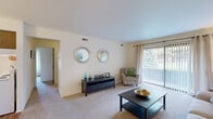 This two-bedroom, one-bathroom residence in Clinton Township, Michigan is thoughtfully laid out across a single floor, offering a comfortable and efficient use of space. The living room serves as the heart of the home, featuring plush carpeting, neutral warm-toned walls, and a large sliding glass door dressed with vertical blinds that floods the room with natural light and provides outdoor views. The kitchen is equipped with ample white cabinetry accented by wood trim, a stainless steel sink, dishwasher, and durable hardwood-style flooring that extends into the adjacent dining area. The dining space comfortably accommodates a small table and chairs, making it ideal for everyday meals. Both bedrooms are carpeted and feature bifold closet doors with wood-grain finishes, offering practical storage. The primary bedroom is generously sized and includes a window that brings in natural light. A connecting hallway ties the rooms together efficiently. Throughout the unit, the neutral color palette and clean lines create a calm, cohesive ambiance suited for comfortable everyday living.