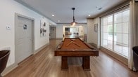 This single-floor games and social lounge at Wesley offers a thoughtfully designed space for recreation and casual gathering. The open layout is anchored by a handsome, full-size billiards table with a rich wood frame and amber felt, positioned beneath a stylish pendant light fixture that adds a warm, inviting glow. Warm hardwood-style flooring runs throughout, tying together the various activity zones seamlessly.

A dedicated game area along one wall features a regulation shuffleboard table, framed by textured accent wall panels and decorative artwork. Multiple bistro-style bar-height tables paired with contemporary swivel stools are distributed throughout the space, providing ample casual seating for socializing. A wall-mounted flat-screen television enhances the lounge experience in the seating alcove.

Large glass doors and generous windows along the perimeter flood the interior with natural light while offering views of lush greenery outside, creating a bright and airy atmosphere. Recessed lighting throughout ensures the space remains well-lit at all hours. An adjacent lounge area, visible through an open archway, extends the social experience with additional comfortable seating. The overall ambiance strikes a balance between energetic recreation and relaxed socializing, making it an ideal amenity space for community use.