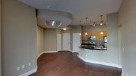 This single-floor residence showcases an open-concept layout that seamlessly connects the living area to the kitchen. A standout architectural feature is the curved tray ceiling in the living area, which incorporates recessed lighting and adds a distinctive sculptural element to the space. The rich, dark hardwood-style flooring runs throughout, grounding the interior with warmth and continuity.

The kitchen is anchored by a curved island with a dark countertop, offering both workspace and a natural visual divider between the cooking and living areas. Warm wood cabinetry lines the kitchen walls, complemented by stainless steel appliances including a range, microwave, and refrigerator. Pendant lights hang above the island, providing focused task lighting while enhancing the overall aesthetic.

Neutral taupe-toned walls create a versatile backdrop throughout the space, while white baseboards and door trim offer clean contrast. Several interior doors are visible along the back wall, indicating additional rooms beyond the main living and kitchen area. Track lighting in the kitchen zone supplements the pendant fixtures, ensuring the workspace is well-illuminated. The overall ambiance is modern, functional, and cohesive.