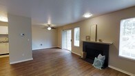 This single-story space centers around a well-appointed living room that blends comfort and functionality. Rich, dark hardwood flooring extends throughout, providing a warm and grounded aesthetic that anchors the room. A classic fireplace with a black surround and white mantle serves as the focal point of the space, offering both visual interest and a cozy gathering spot. Adjacent to the fireplace, French-style sliding glass doors are flanked by a side window fitted with blinds, allowing natural light to filter into the room while providing access to the outdoors. A ceiling fan with a decorative light fixture is centrally positioned, ensuring year-round comfort and adding to the room's practical design. The walls are finished in a neutral beige tone, complementing the warm flooring and creating a versatile backdrop. A textured popcorn ceiling adds a traditional touch overhead. Flush-mounted lighting provides additional illumination, while a wall vent indicates an integrated climate control system. The overall ambiance is inviting and functional, with a layout that offers generous open space throughout the living area.