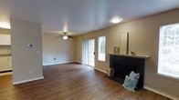 This single-story space centers around a well-appointed living room that blends comfort and functionality. Rich, dark hardwood flooring extends throughout, providing a warm and grounded aesthetic that anchors the room. A classic fireplace with a black surround and white mantle serves as the focal point of the space, offering both visual interest and a cozy gathering spot. Adjacent to the fireplace, French-style sliding glass doors are flanked by a side window fitted with blinds, allowing natural light to filter into the room while providing access to the outdoors. A ceiling fan with a decorative light fixture is centrally positioned, ensuring year-round comfort and adding to the room's practical design. The walls are finished in a neutral beige tone, complementing the warm flooring and creating a versatile backdrop. A textured popcorn ceiling adds a traditional touch overhead. Flush-mounted lighting provides additional illumination, while a wall vent indicates an integrated climate control system. The overall ambiance is inviting and functional, with a layout that offers generous open space throughout the living area.