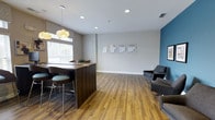 Located at 203 Magnolia Blvd in Port Wentworth, Georgia, this single-floor commercial space offers a clean, modern environment designed for professional use. The open-concept layout is anchored by a dark walnut-finished reception or bar-height counter, accented by stylish pendant lighting with a distinctive floral design, creating an inviting focal point upon entry. The flooring throughout features warm honey-toned hardwood planks that lend a sense of continuity and warmth across the space. Recessed lighting in the ceiling provides consistent, even illumination, complemented by natural light streaming in through window treatments along the exterior wall. The main area includes a comfortable seating arrangement with contemporary grey upholstered chairs, ideal for waiting or informal meetings. An accent wall in deep teal blue adds a bold design element, contrasting tastefully with the neutral grey and white tones that define the rest of the interior. Informational materials are displayed along one wall, suggesting active use as a leasing office or sales center. The overall ambiance is professional yet welcoming, with thoughtful design details that balance function and aesthetics in a well-maintained, move-in-ready environment.