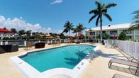 This inviting waterfront property showcases a resort-style outdoor setting designed for relaxation and leisure. At its centerpiece is a generously sized swimming pool with crystal-clear turquoise water, accessible via stainless steel handrails and surrounded by a wide, light-toned stone pool deck. The deck provides ample space for lounge chairs and outdoor seating, making it well-suited for sunbathing or casual gatherings.

Mature palm trees and lush tropical greenery frame the space, lending it a distinctly Florida atmosphere. A shaded seating area with additional patio furniture and a red umbrella offers a comfortable retreat from the sun. The property overlooks a calm canal, with boats visible along the waterway, enhancing the outdoor ambiance with tranquil water views.

In the background, a two-story yellow building with a staircase and covered balcony area is visible, adding to the property's layered, resort-like character. The combination of vibrant landscaping, waterfront access, and thoughtfully arranged outdoor amenities creates a welcoming environment ideal for warm-weather enjoyment under open blue skies.