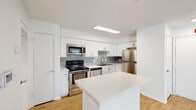 This single-floor residence offers a clean, modern aesthetic throughout its well-designed layout. The kitchen serves as a functional centerpiece, featuring white shaker-style cabinetry, light quartz countertops, and a gray subway tile backsplash. A full suite of stainless steel appliances—including a range, microwave, dishwasher, and refrigerator—completes the space, while a central island provides additional prep and seating space. Light wood-look flooring flows seamlessly from the kitchen into the open living area.

The living room is bright and airy, with natural light streaming through large windows dressed with horizontal blinds. A ceiling fan overhead and a fireplace with a clean white surround add both comfort and character to the space. The open sightlines between the kitchen and living area enhance the sense of spaciousness.

The bedroom is carpeted for warmth and comfort, features a ceiling fan, and receives natural light through a window overlooking greenery. The bathroom is finished with white cabinetry, a quartz-topped vanity, a large mirror, and a tub-shower combination. A walk-in closet with built-in shelving is accessible directly from the bathroom, offering practical storage solutions throughout.