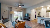 This single-floor residence offers a thoughtfully designed open-concept layout that seamlessly connects the living, dining, and kitchen areas. The living room is anchored by wood-look flooring, a ceiling fan, and a bold teal accent wall, with a sliding glass door that opens to a private patio overlooking a lush green outdoor area, filling the space with natural light. The kitchen is a standout feature, showcasing rich dark cabinetry, granite-style countertops, a subway tile backsplash, stainless steel appliances including a refrigerator, range, microwave, and dishwasher, and elegant pendant lighting above a peninsula with a sink. The dining area provides an intimate setting with its own accent wall detail and decorative touches. The bedroom is a comfortable retreat, highlighted by plush carpet, a patterned feature wall, large windows with blinds, and warm ambient lighting. The bathroom is finished with granite countertops, dark wood-tone cabinetry, a large framed mirror, and a full shower-tub combination. Throughout the residence, consistent wood-look flooring, neutral tones, and modern fixtures create a cohesive and inviting atmosphere suited for contemporary living.